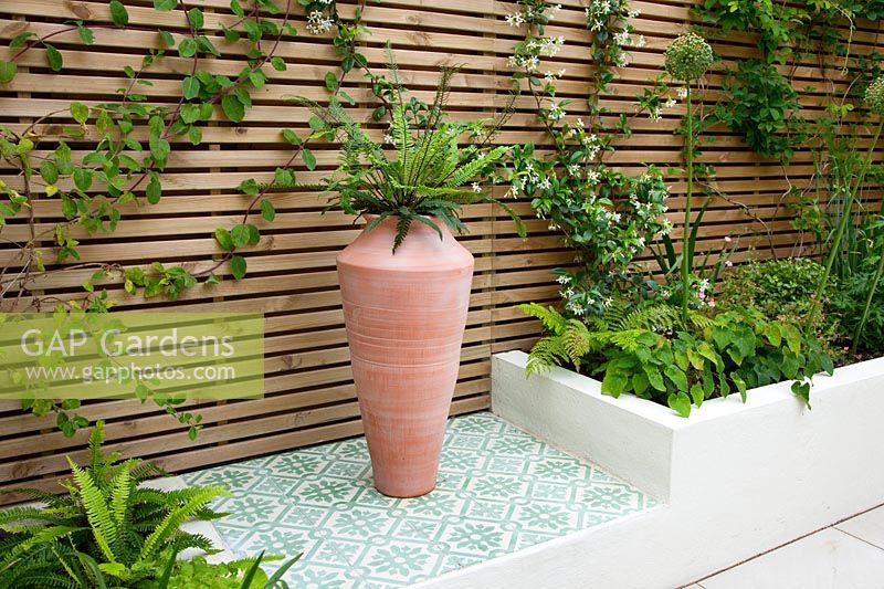Plinth paved with morroccan tiles and raised beds of ferns, with large oil jar and slated trellis, June