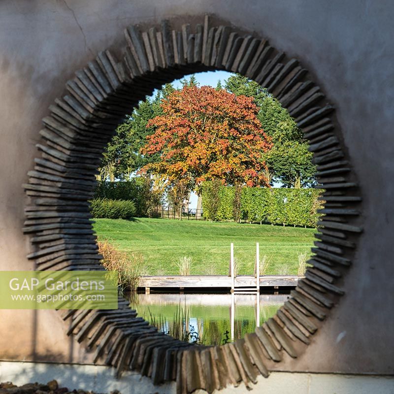View through moongate to deck of natural swimming pool with autumn foliage of a copper sycamore.