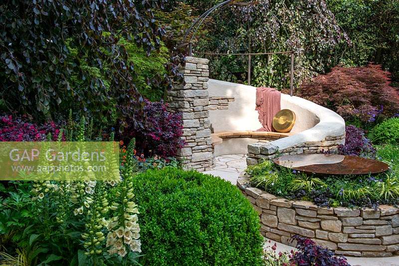 The Kingston Maurward Garden at RHS Chelsea Flower Show 2019. A curving seating area in Purbeck stone, surrounded by purple beeches, acers and other herbaceous planting. Design: Michelle Brown. Sponsors: Miles Brown, Kingston Maurward College, Goulds Garden Centre, Wilks Landscaping, Holme for Gardens, The Green Gardener.
