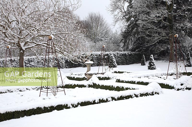 View of snow-covered, formal garden, featuring low Buxus sempervirens - Box - beds with decorative iron obelisks, a central sundial and Taxus baccata - Yew - cones.
