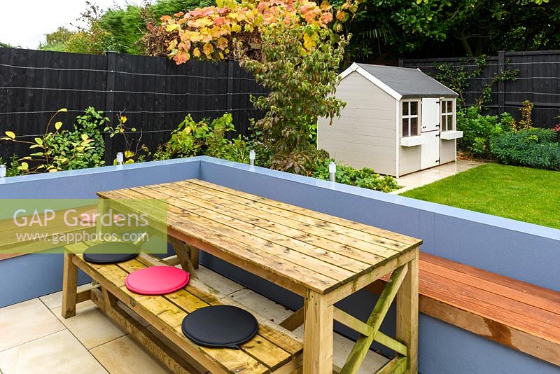 Wooden table and bench dining area look out to child's playhouse in modern garden. 