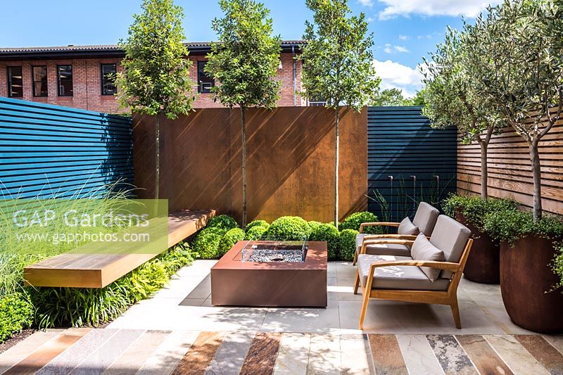 Contemporary seating area with fireplace surrounded by Olea europaea and
 Muehlenbeckia complexa in rusted steel containers, Quercus ilex, Buxus 
sempervirens balls topiary by rusted panel wall and Miscanthus sinensis
 'Morning Light' by the bench. 