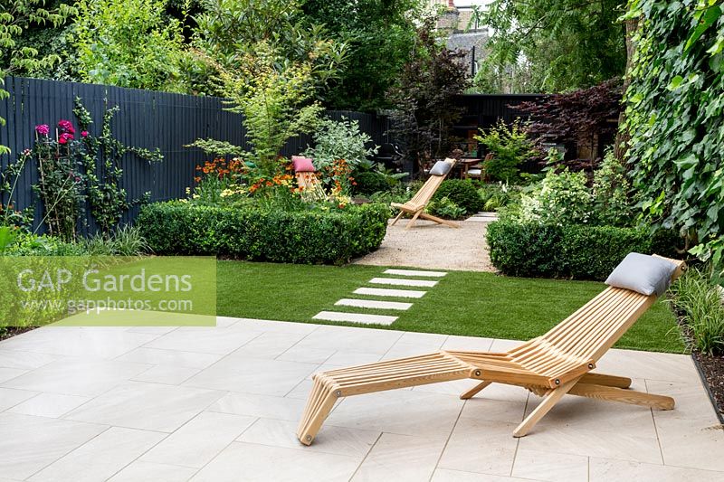 Garden design by Nick Gough 
Overview of garden with artificial lawn with stone slab path and patio with 
wooden chairs. Planting on the left: buxus sempervirens hedge 
Orange Helenium 'Moerheim Beauty' 
Yellow Achillea 'Teracotta'