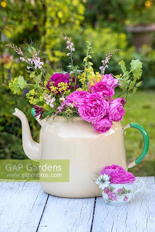 Cut roses arranged in ceramic teapot, against a garden backdrop.
