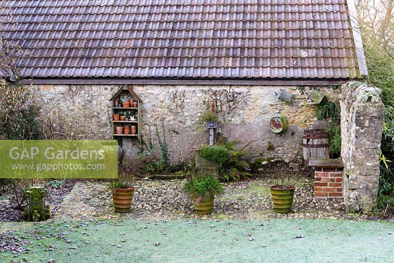 Terracotta pots, galvanised cans and baths and a shelf of decorative items are displayed against a barn. 