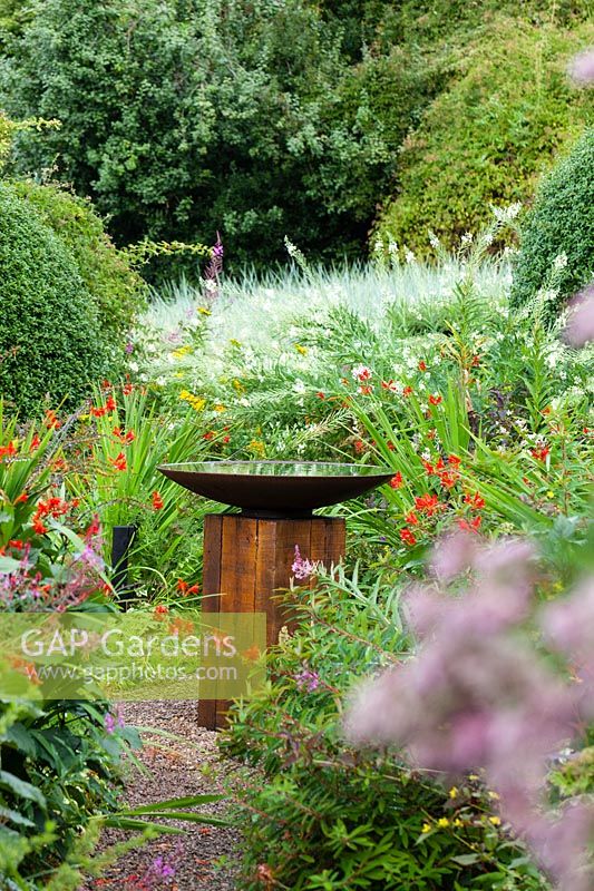 Bird bath of oak sleeper pedestal and mild steel bowl surrounded by Crocosmia 'Lucifer and other perennials at  Veddw House Garden, Monmouthshire, Wales, UK.