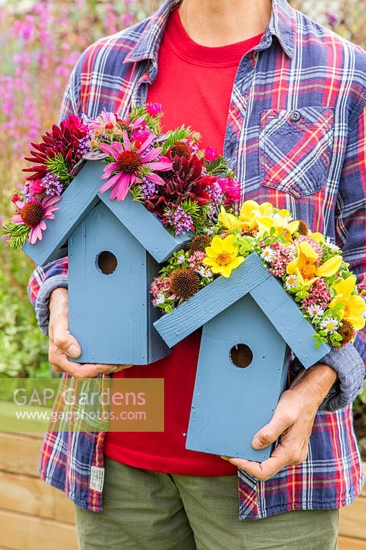 Person holding pair of wooden birdboxes with flower embellished roofs