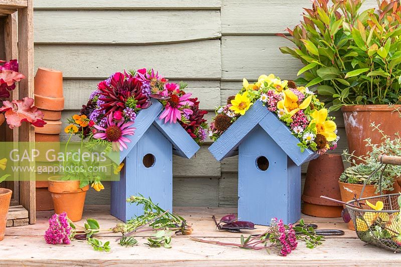 Pair of wooden birdboxes with flower embellished roofs with more cut flower heads on bench with terracotta pots