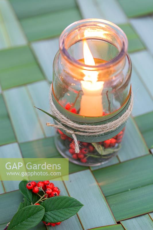 Glass candle holder decorated with Cotoneaster berries, sits on woven Phormium table mat.
