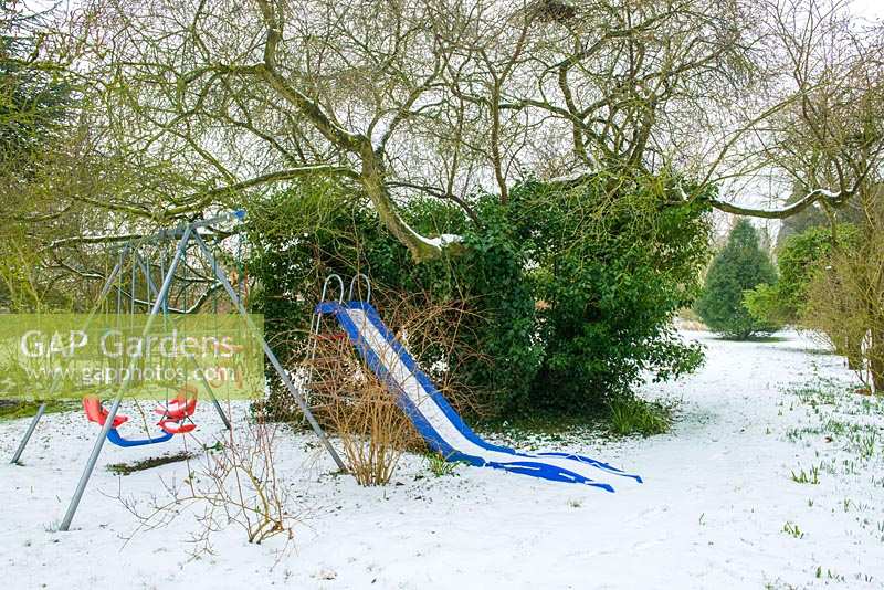 Children's swing and slide set under trees in wild garden with snow. 