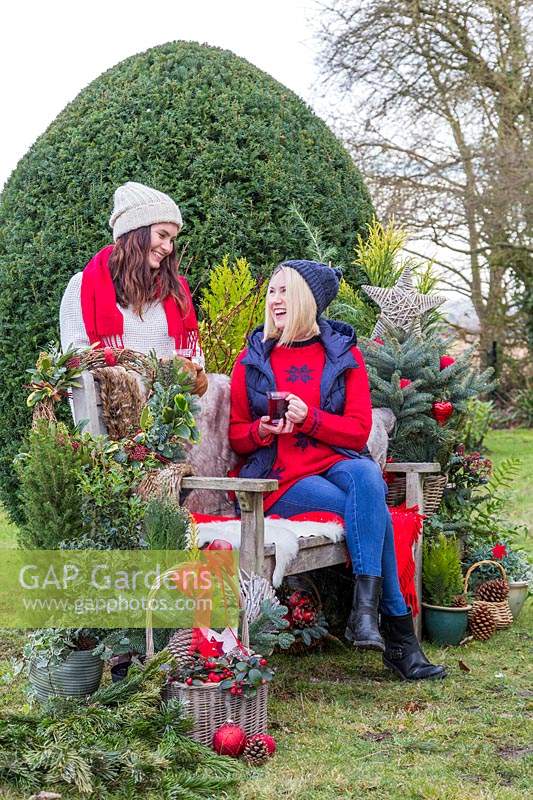Women chatting by bench while having a warm drink, surrounded by Christmas greenery and decorations