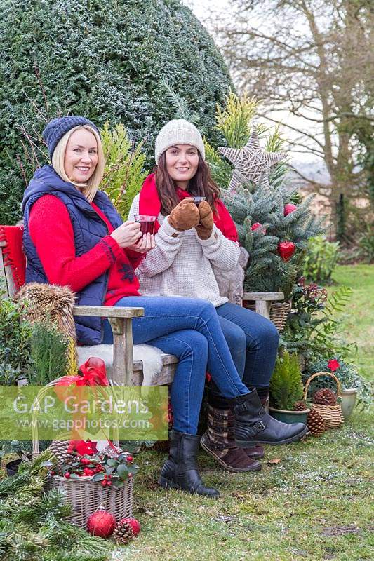 Women chatting by bench while having a warm drink, surrounded by Christmas greenery and decorations