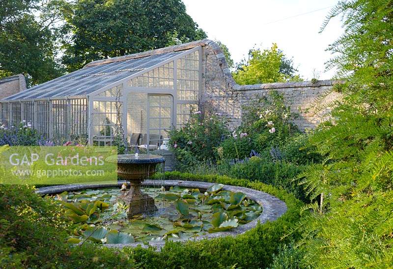 Nymphaea waterlilies in a circular pond with stone fountain surrounded by Buxus - box hedging. Rare Lean-to greenhouse.  Rosa 'Queen of Sweden' and Nepeta grandiflora 'Summer Magic' in front of greenhouse 