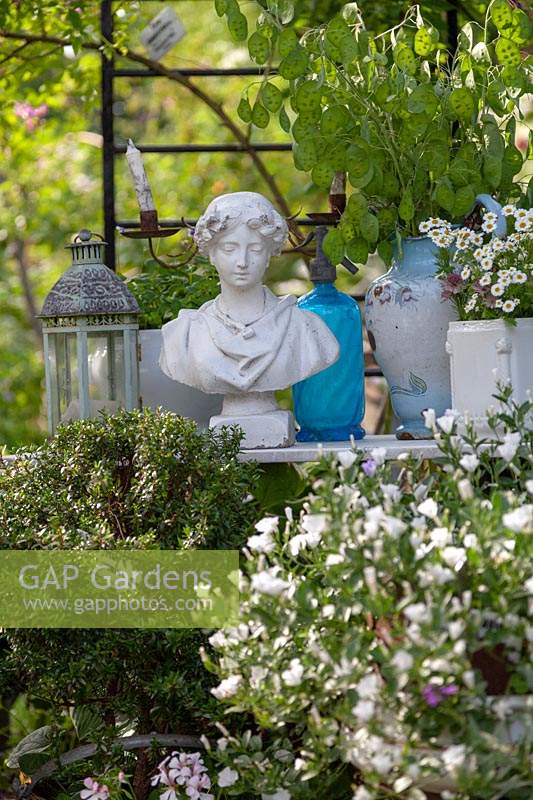 View of table of collectable in a garden, items include a woman's bust 
surrounded by lantern, jug of honesty and potted plant