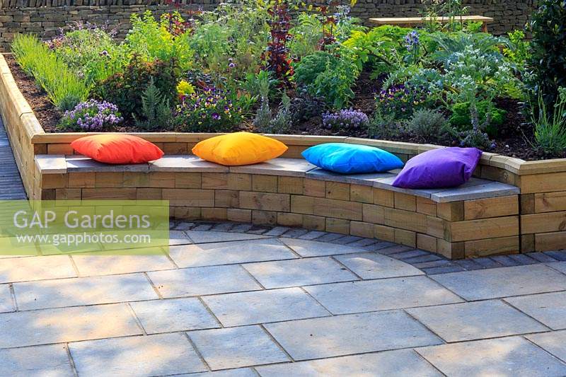Brightly-coloured cushions add colour to the central circular sitting area, with herbs growing in raised bed behind. 'Health and Wellbeing' permanent garden designed by Jekka McVicar, Malvern Spring Festival, UK. 