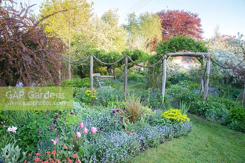View across packed flower beds separated by a decorative rope feature and 
rustic archway. Planting in foreground includes: Berberis x ottawensis, Tulipa, 
Camassia, Geum, Euphorbia, Centaurea montana, and Myosotis.
