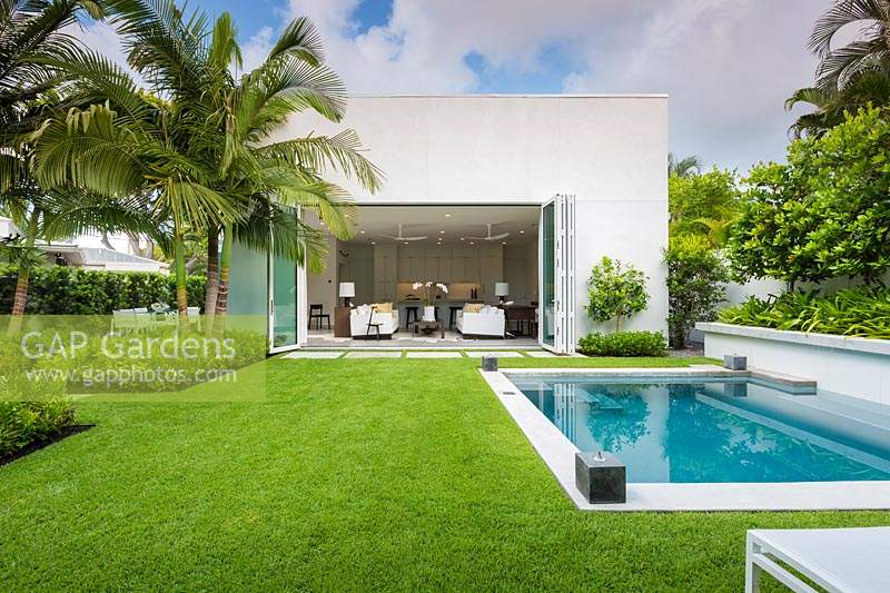 View over Zoysia grass lawn to back of house with folding doors, on one side a swimming pool and on the other beds of foliage plants including palms