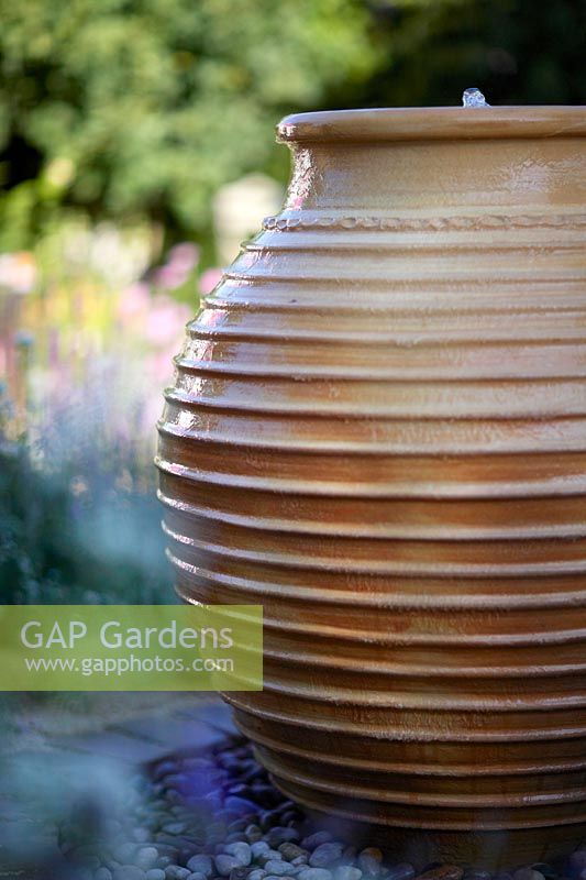 Urn-shaped terracotta water feature on pebbles. Best of Both Worlds Garden, Sponsored by BALI, RHS Hampton Court Palace Flower Show, 2018.
