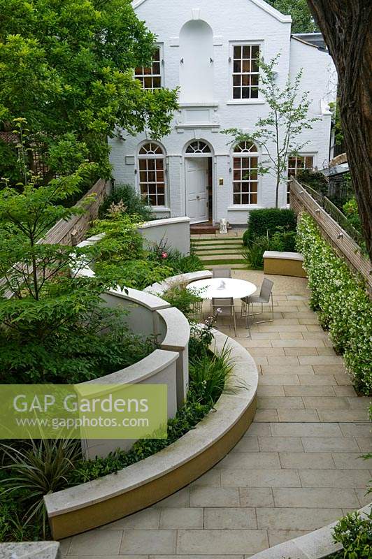 View of private urban back garden and house, with pale paving and curved bench and wall. 