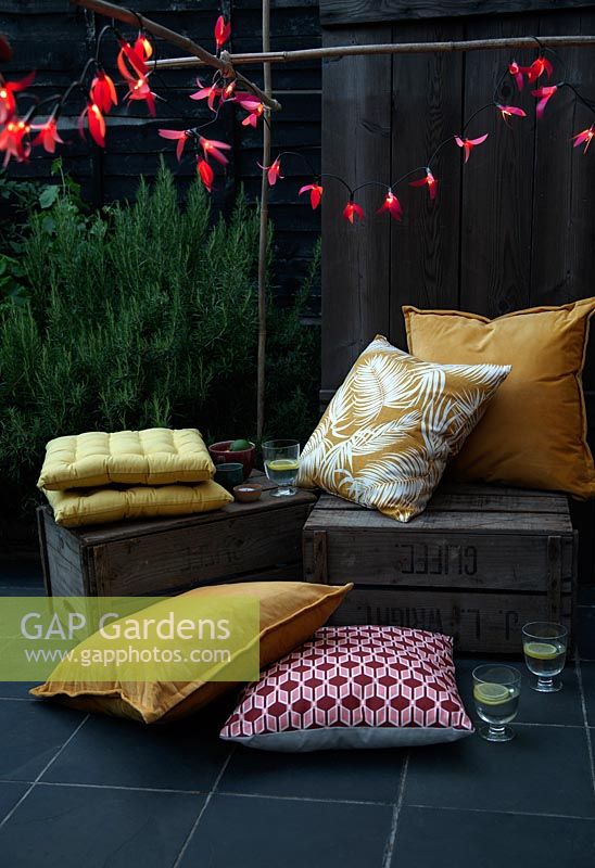 Fairylights decorated with colourful paper shapes to create beautiful decorative outdoor lighting, used here over bamboo cane supports in a seating area with fruit crates and cushions