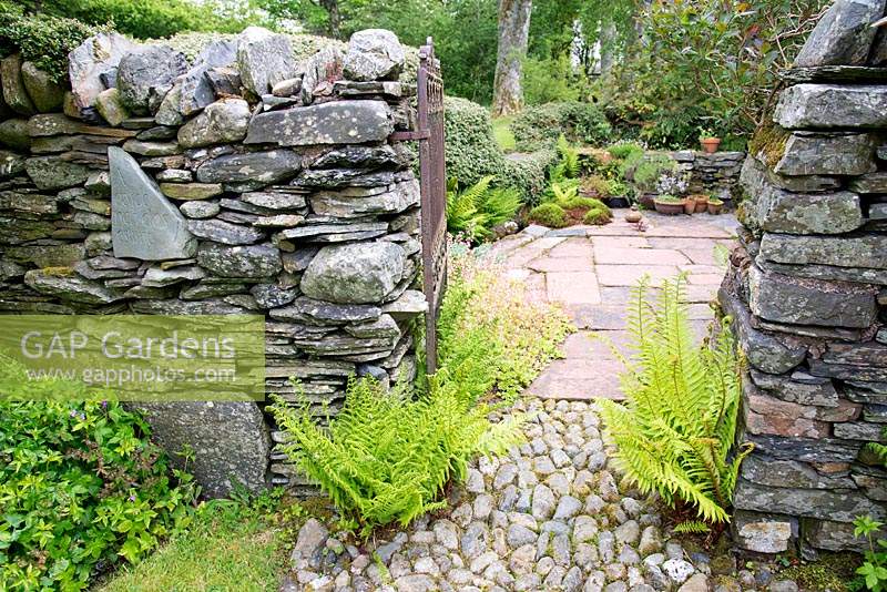 Stone wall with inscribed slate, and gate which leads to paved area with containers. 
