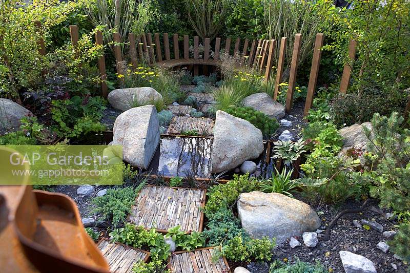 Through Your Eyes Garden. Wooden seating area with slate pieces inlaid into corten steel frames to form stepping stones. Water pool with 'split' rock.   Planting includes Deschampsia cespitosa, Achillea 'Moonshine', Ophiopogon planiscapus 'Nigrescens' - Sponsors: Kebony, CED Stone, R and G Metal Products, William's Art and Design, Practicality Brown.