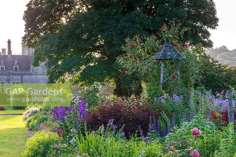 A section of a large deep border packed with plants and featuring an obelisk with climbing red Rosa - Rose