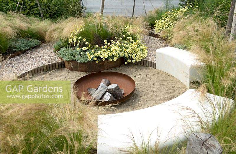 Fireplace with wood and inscription BONFEU. Stipa tenuissima and Anthemis x hybrida planted in border with rust edges. Path with shells. Cosy round seating area with benches of stone.