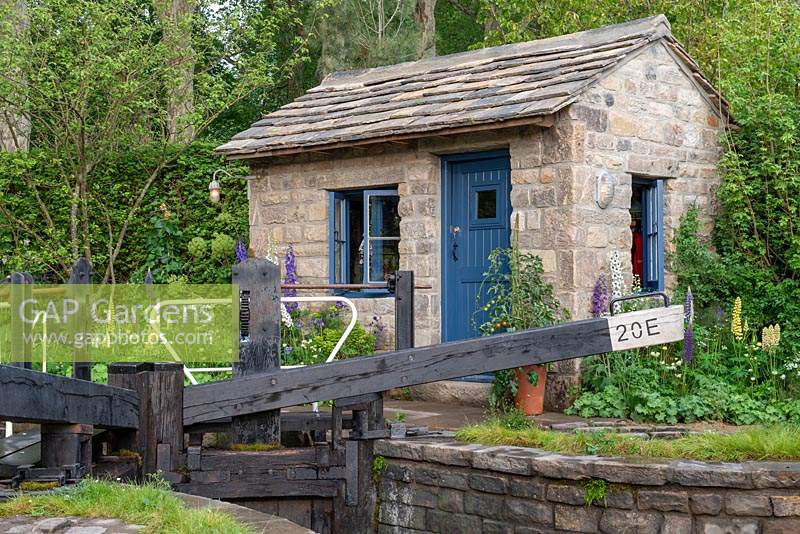 Lock keeper's office next to the lock gates with herbaceous planting of Lupins and Delphiniums - The Welcome to Yorkshire Garden, RHS Chelsea Flower Show 2019.