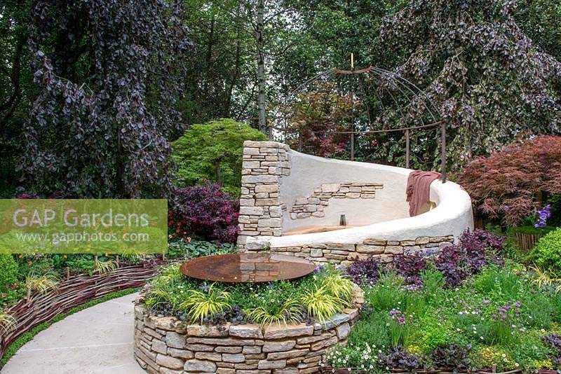 Fagus sylvatica 'Purple Mountain' trees providing the backdrop to a dry stone wall seating area with decorative sundial dome and circular pool, plants include Loropetalum, Acer palmatum, Carex and Erigeron karvinskianus - Miles Stone: The Kingston Maurward Garden, RHS Chelsea Flower Show 2019.