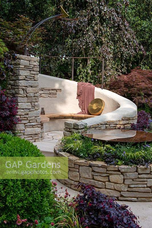 Fagus sylvatica 'Purple Mountain' trees providing the backdrop to a dry stone wall seating area with decorative sundial dome and circular pool, plants include Loropetalum, Acer palmatum and Carex - Miles Stone: The Kingston Maurward Garden, RHS Chelsea Flower Show 2019