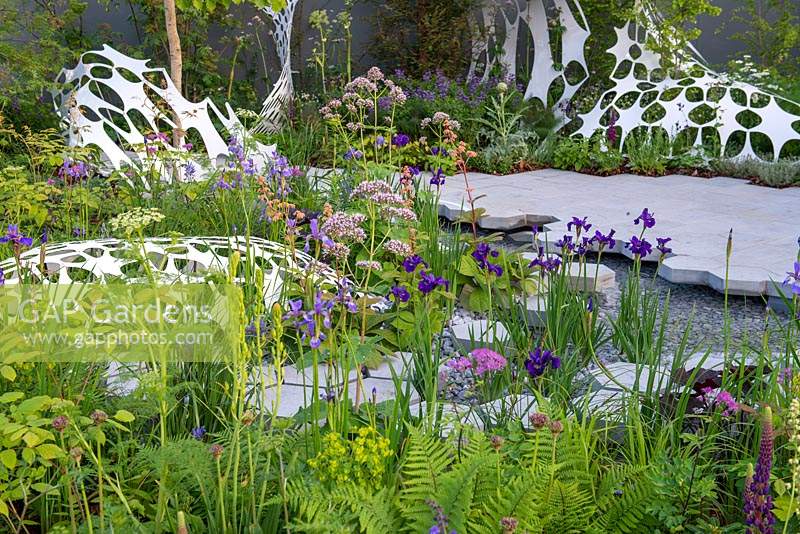 Hexagonal paving next to a pond with Iris sibirica 'Caesar's Brother', ferns and sculpture by Liam Hopkins - The Manchester Garden, RHS Chelsea Flower Show 2019.
