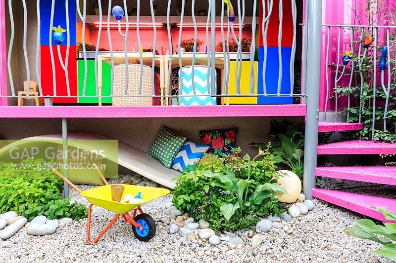 The Montessori Centenary Children's Garden. Detail of crawl space under bright pink children's educational playroom. Child's yellow wheelbarrow in the foreground, pink spiral staircase to one side. Plants include various Mentha cvs - Mint, Astilbe and Hosta 'Devon Green'. Sponsor:  Montessori Centre International