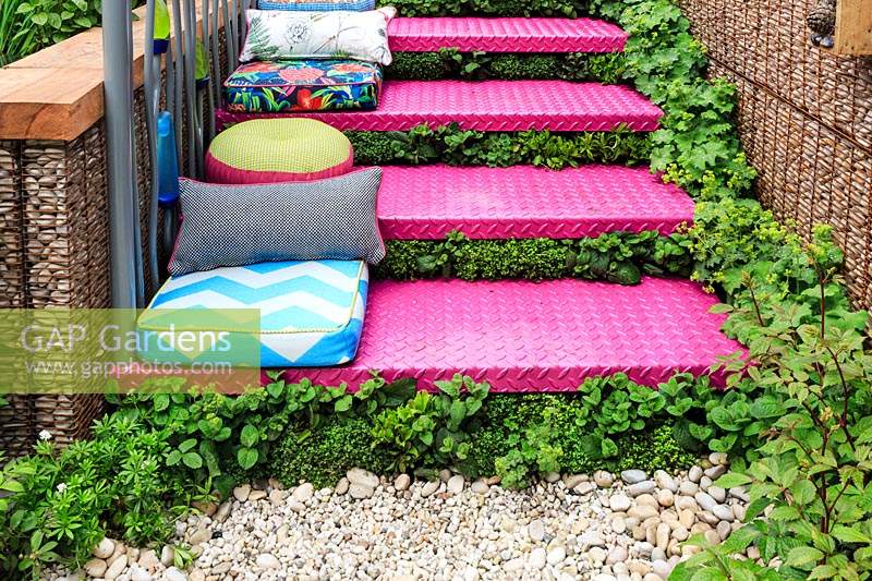 The Montessori Centenary Children's Garden. Detail of planting in between bright pink metal steps with brightly patterened and coloured cushions for children to sit on. Planting includes: Mentha - Mint, Soleirolia soleirolii and Alchemilla mollis. Sponsor:  Montessori Centre International