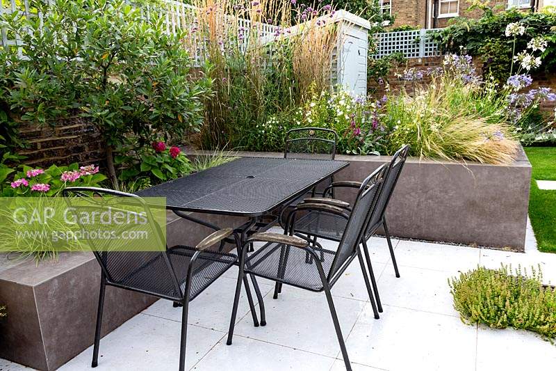 Black metal table and chairs on white paved patio. Grey raised beds with planting including: Gaura lindheimeri 'Karalee White', Agapanthus Blue Heaven, Agapanthus 'Queen Mum', Calamagrostis 'Karl Foerster', Laurus nobilis - Bay