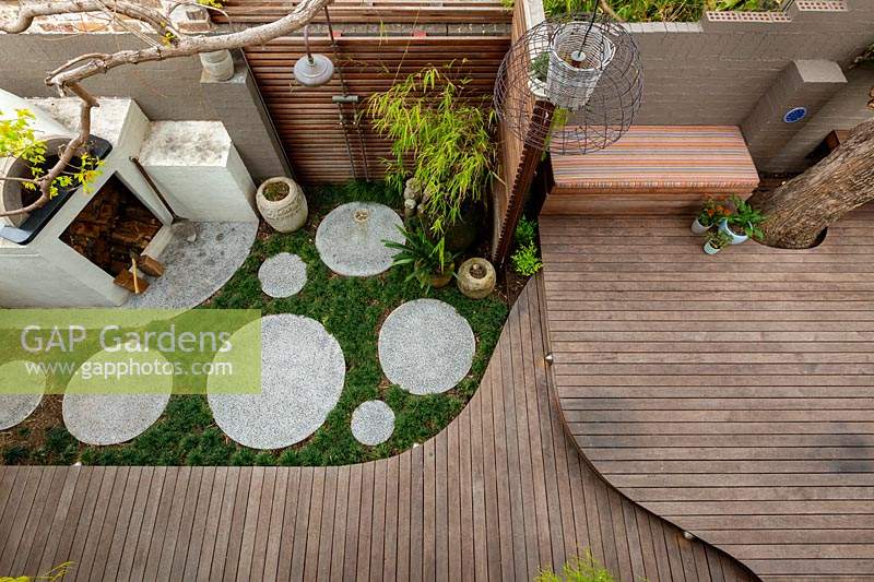 Overhead view of a backyard, from a first floor balcony showing a bespoke cement rendered pizza oven in the back corner of a backyard showing a slat hardwood timber screen, a timber deck, a clump of bamboo, groups of pots and a Jacaranda tree growing through the deck.