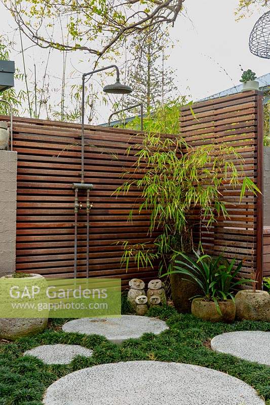 Corner of a garden next to a painted brick fence, featuring a timber slat screen with an outdoor shower, a stone carved Indonesia pot, a family group of sculptures, a large glazed water bowl, semi rustic stone pots, concrete circular cast in situ stepping stones in amongst a thick planting of mini Mondo grass.