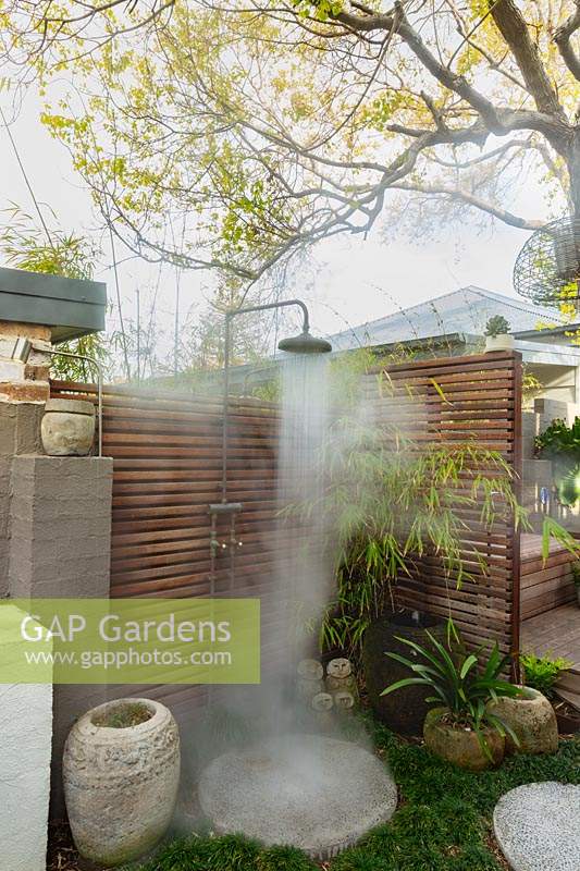 An outdoor shower, showing hot water coming out of it, attached to a hardwood timber privacy screen, with potted bamboo, pots and round stepping stones.