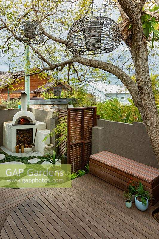 A wide view of a backyard, with a bespoke cement rendered pizza oven in the back corner of a backyard showing a slat hardwood timber screen, a timber deck, a clump of bamboo, groups of pots and a Jacaranda tree growing through the deck.