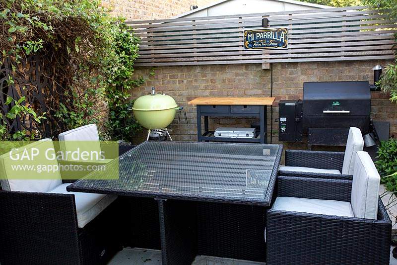 Seating and dining patio area in small shade tolerant garden in London with a green theme. View across the patio towards table and barbecue.