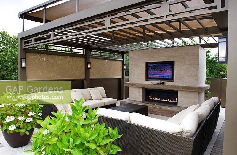 View of outdoor room on a roof garden, showing relaxation area of sofas, flat screen television and outdoor fire pit. All sheltered by metal pergola with wall lights 