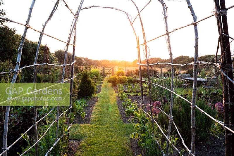 Grass path through the vegetable garden framed by a simple hazel archway which will be covered with sweet peas later in the season at Sea View, Cornwall, UK in June.
