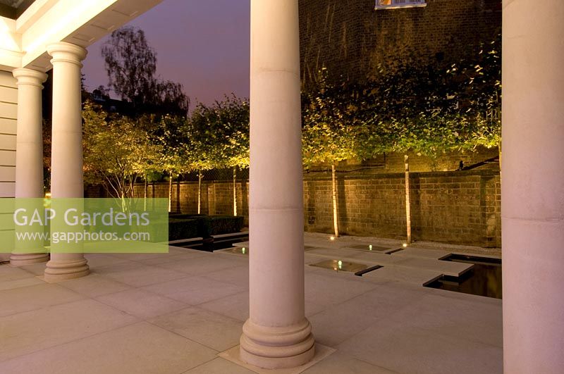View through white paved terrace with pillars to a row of standard trees, lit up with spotlights in contemporary, city garden. 