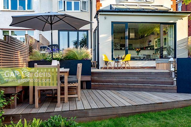 Contemporary garden with split level decking. The lower section includes a bespoke built in bench with cushions, a table with parasol and chairs, alongside grey raised beds with perennials.