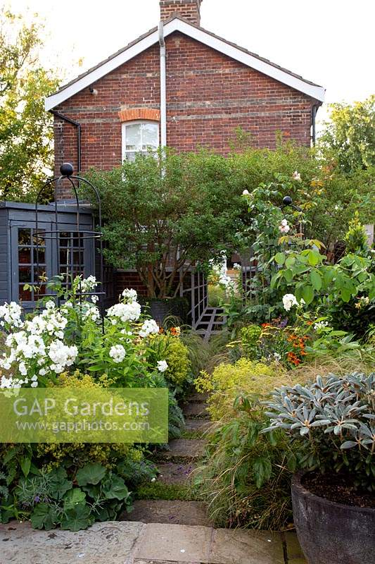 Small garden, view from paved patio along path with flower beds either side. Planting includes: Phlox 'David', Achillea 'Moonshine', Stipa arundinacea and Alchemilla mollis. Other features include: painted summerhouse, obelisk, tree in large pot and trompe l'oeil of mirror and trellis
