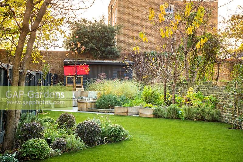 View across the garden with artificial grass to seating area with fire pit and round wooden bench surrounded by Miscanthus sinensis 'Morning Light' 'eulalia', Prunus avium 'Stella', Rubus idaeus, Pittosporum tenuifolium 'Tom Thumb' and raised vegetable beds 