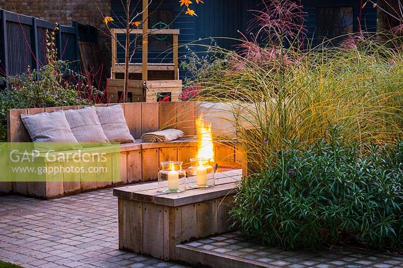 Seating area with fire pit and round wooden bench surrounded by Miscanthus sinensis 'Morning Light' - eulalia and Erysimum 'Bowles's Mauve' 