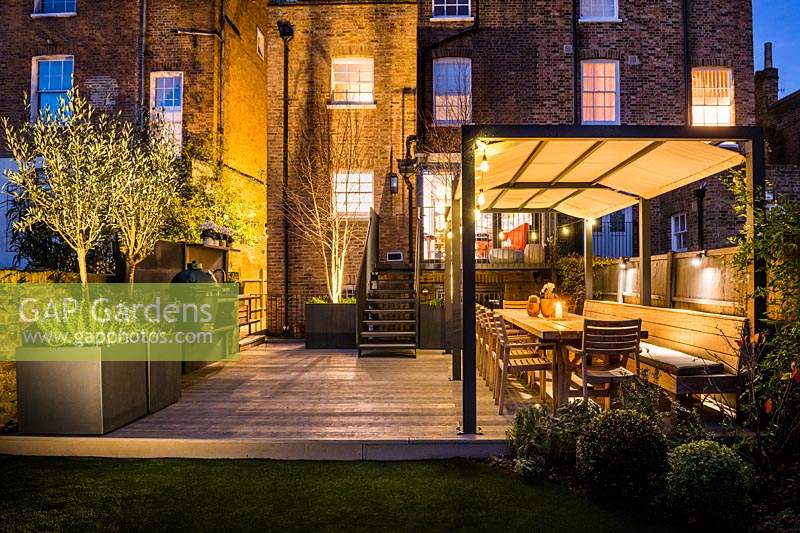 Illuminated outdoor kitchen and dining area with wooden tables and chairs and plants in containers including Olea europaea at night. 