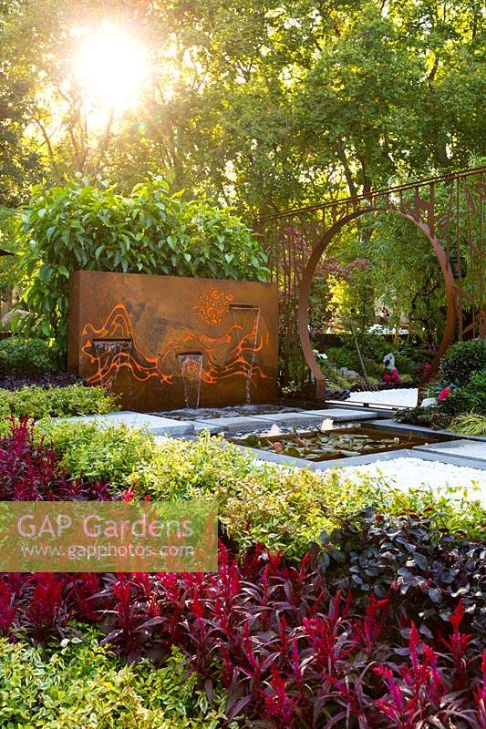 Pavilion style Chinese garden, with a freestanding water feature, moongate, stepping stones, striped paving , limestone rock garden feature, planter boxes, stone mountain range, printed glass panels, steps and red lanterns.
