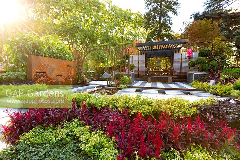 Pavilion style Chinese garden, with a freestanding water feature, moongate, stepping stones, striped paving, planter boxes, stone mountain range, printed glass panels, steps and red lanterns.